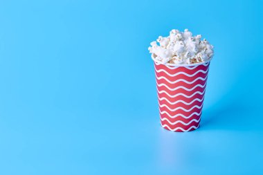 Sağ taraftaki mavi arka planda patlamış mısır dolu kırmızı bir bardak duruyor. Boş alan. Film ve TV programları için yemek konsepti.