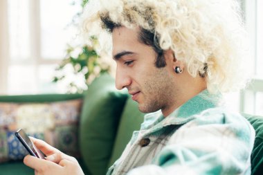 Evdeki kanepede oturan genç adam, cep telefonu kullanıyor.