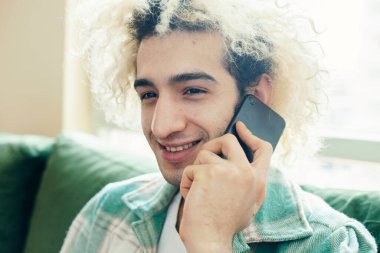 Evdeki kanepede oturan genç adam, cep telefonu kullanıyor.