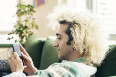 Evdeki kanepede oturan genç adam, cep telefonu kullanıyor.