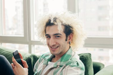 Evdeki kanepede oturan genç adam, cep telefonu kullanıyor.