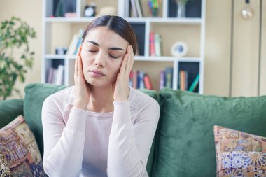 Üzgün depresif kadın evinde, koltukta oturuyor, aşağıya bakıyor ve alnına dokunuyor, yalnızlık ve acı kavramı.