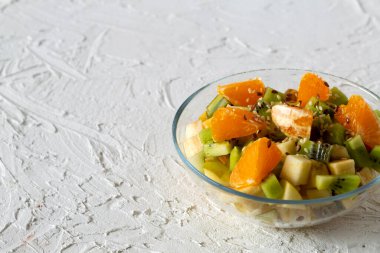 Sağlıklı vejetaryen yemeği. Beyaz arka planda cam kasede meyve salatası. Beyaz bir masada yeşil naneli meyveli bir kase sağlıklı salata. Karışık taze muz, portakal, kivi, meyve. Boşluğu kopyala.