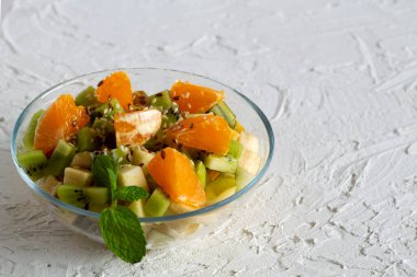 Sağlıklı vejetaryen yemeği. Beyaz arka planda cam kasede meyve salatası. Beyaz bir masada yeşil naneli meyveli bir kase sağlıklı salata. Karışık taze muz, portakal, kivi, meyve. Boşluğu kopyala.