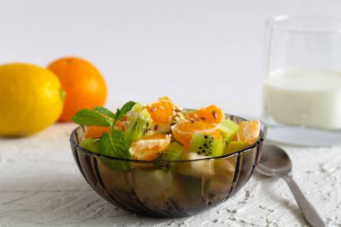Beyaz arka planda bir bardak sütle cam kase içinde meyve salatası. Beyaz bir masada yeşil naneli meyveli bir kase sağlıklı salata. Karışık taze muz, portakal, kivi, meyve. Boşluğu kopyala.