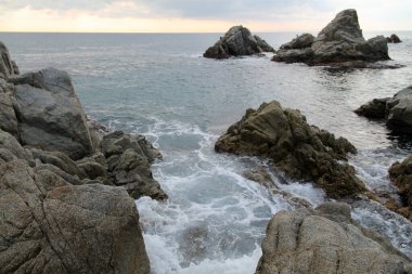 Denizin üzerinde gün batımı. Akdeniz manzarası çok güzel. Sahilde büyük taşlar, sakin su ve bulutlu arka plan