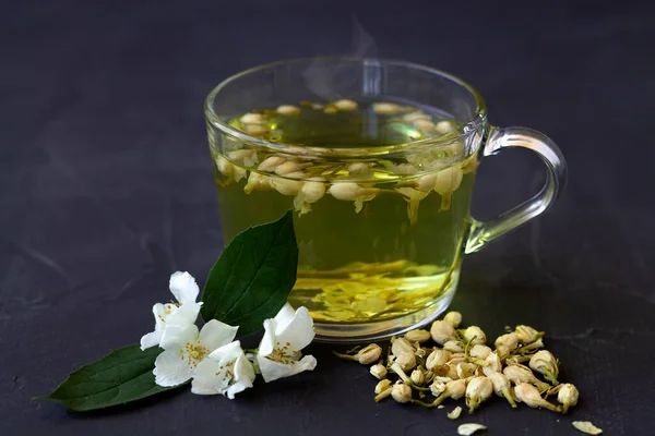 Close up Jasmine tea. Cup of hot herbal tea with jasmine fresh flowers on a black table. Healthy lifestyle. glass cup of green tea with dry leaves on a black background. Hot drinks.