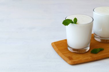 Beyaz masada yeşil naneli iki bardak yoğurt. Taze süt içeceği. Fotokopi alanı ile mayalanmış sağlıklı ürünler konsepti. Kefir, süt ya da Lassi masada probiyotiklerle birlikte içerler. Hint mutfağı.