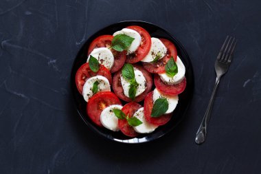 Siyah tabakta çatal ve fotokopi alanı olan İtalyan caprese salatası. Dilimlenmiş kırmızı domates, mozzarella peyniri, yeşil fesleğen, koyu arkaplanda zeytinyağı. Taze salata. Üst manzara..