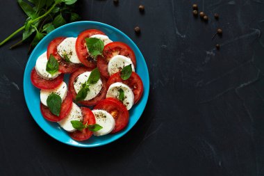 Domates ve mozzarella. İtalyan caprese salatası. Mavi tabakta yeşil ve fotokopi alanı var. Dilimlenmiş kırmızı domates, mozarella peyniri, yeşil fesleğen, siyah arka planda zeytinyağı. Taze salata. Üst manzara..