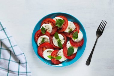 Mavi tabakta çatallı taze salata. Geleneksel İtalyan Caprese salatası. Dilimlenmiş kırmızı domates, mozzarella peyniri, yeşil fesleğen, beyaz arka planda fotokopi alanı olan zeytinyağı. Üst görünüm. Düz.