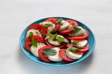 Mavi tabakta İtalyan Caprese salatası. Dilimlenmiş domates, mozarella peyniri, yeşil fesleğen, beyaz arka planda zeytinyağı. Kopya alanı olan taze salata.