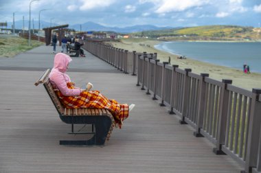 Donmuş genç bir kadın, bir bardak kahve ve cep telefonuyla güneşlenip kareli battaniyeyle örtülmüş. Kumsalda soğuk bir sonbahar günü