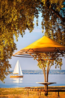Körfezde ağaç dallarıyla çevrili bir çardak ve ufukta bir yelkenli yatı var. Güneşli bir günde sonbahar manzarası.