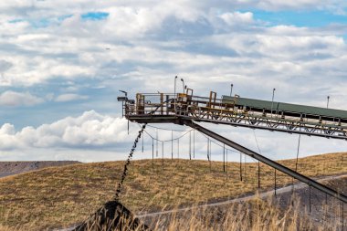 Uzun bir endüstriyel taşıyıcının altında büyük bir kömür deposu var. Sahne, Avustralya kömür endüstrisinin engebeli ve işlevsel manzarasını vurgulayarak, madencilik operasyonları ve çıkarma sürecinin ölçeğini yakalıyor..