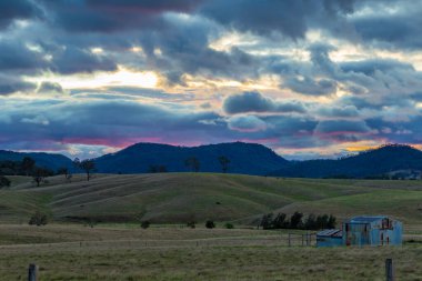 Dramatik bulutlu bir gökyüzü altında uzak dağlara doğru uzanan açık tarım arazilerinin olduğu manzaralı bir kırsal bölge. Görüntü, kırsal kesimin doğal güzelliğini ve huzurlu atmosferini yansıtıyor. Yumuşak ışık dokuyu aydınlatıyor..
