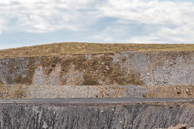 Engebeli bir maden yolu, büyük kuyruk yığınlarıyla çevrili bir endüstriyel maden sahasından geçiyor. Sahne, mineral çıkarmanın ölçeğini ve etkisini, sanayi altyapısını, toprak çalışmalarını ve madencilik arazilerinin ham dokularını gösteriyor.