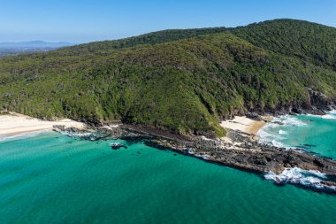 Forster 'daki Seven Mile Plajı' nda Janies Corner 'ı çeken hava görüntüsü geniş bir kumla buluşan okyanus dalgalarını gösteriyor. Yükseltilmiş perspektif kıyı dokularını, sörf hatlarını ve berrak mavi suyu vurgular ve manzaralı bir deniz manzarası yaratır..
