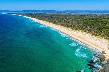 Forster 'daki Seven Mile Plajı' nda Janies Corner 'ı çeken hava görüntüsü geniş bir kumla buluşan okyanus dalgalarını gösteriyor. Yükseltilmiş perspektif kıyı dokularını, sörf hatlarını ve berrak mavi suyu vurgular ve manzaralı bir deniz manzarası yaratır..