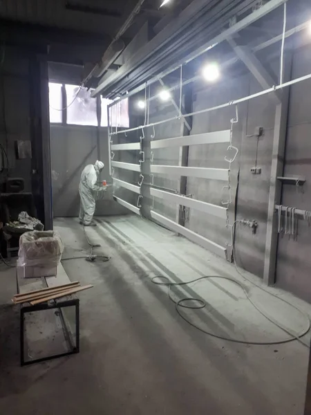 A male painter in a white disposable protective suit paints a metal structure with a spray gun at a paint factory.