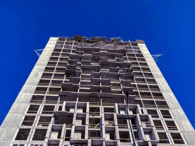 Büyük, tamamlanmamış beton bir bina ve bir inşaat alanında mavi gökyüzüne karşı yeni binalar..