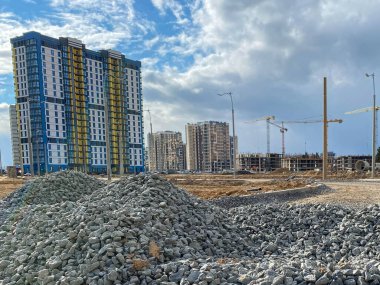 Büyük gri taşlar, endüstriyel yol inşaatından kalma molozlar ve şantiyede vinçleri olan yeni binalar..
