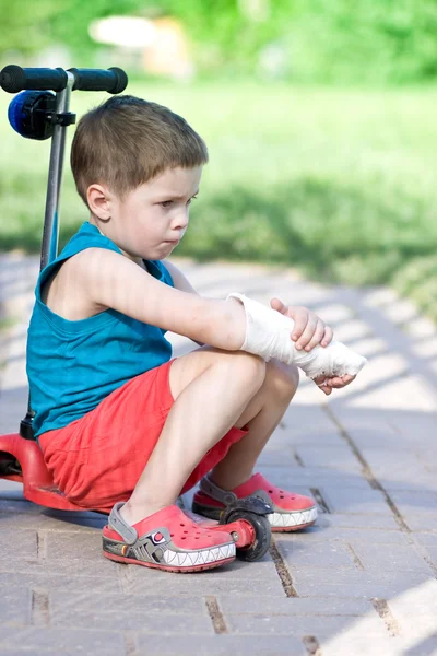 Triste chico con el brazo roto se sienta en scooter — Foto de Stock