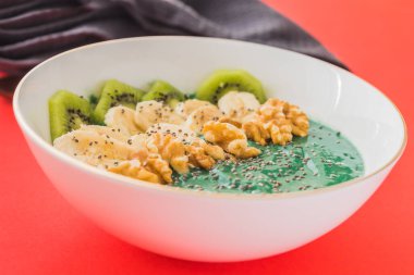 Kırmızı zemin üzerinde muz, ceviz, kivi ve chia tohumları olan bir spirulina smoothie kasesi.