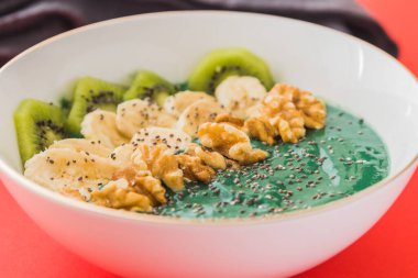 Kırmızı zemin üzerinde muz, ceviz, kivi ve chia tohumları olan bir spirulina smoothie kasesi.