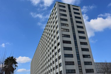 Parlak mavi gökyüzü ve dağınık beyaz bulutlar altında birçok penceresi olan yüksek şehir binası. Aşağıdan çekilen modern konut veya ofis yapısı temiz çizgiler ve geometrik simetri yakalıyor.