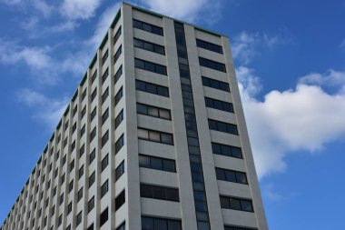 Parlak mavi gökyüzü ve dağınık beyaz bulutlar altında birçok penceresi olan yüksek şehir binası. Aşağıdan çekilen modern konut veya ofis yapısı temiz çizgiler ve geometrik simetri yakalıyor.