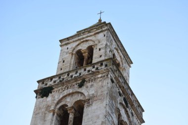 Casamassima 'daki tarihi çan kulesinin manzarası, yaşlandırılmış taş mimari, kemerli açıklıklar ve tepesinde bir haç var. Duvarın ılık tonlarıyla temiz gökyüzü, eski kasabanın mirasını ve çekiciliğini vurguluyor..
