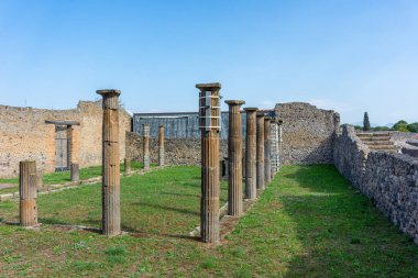 Harabeler ve duvarlarla çevrili çimenli bir arkeolojik alan boyunca sıralanmış taş sütunları olan antik bir Roma sütununun görüntüsü. Sahne klasik mimariyi, şehir tasarımını ve bir Roma şehrinin korunmuş yapısını yansıtıyor..