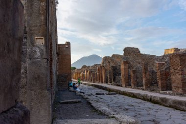 Arka planda Vesuvius Dağı ve harabeleri keşfeden ziyaretçilerin bulunduğu Pompeii, İtalya 'nın arkeolojik sahasında antik bir Roma kaldırım caddesinin görüntüsü. İkonik tarihi ve kültürel sahne.