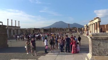 Zaman atlaması, Pompei 'nin arkeolojik harabelerinde büyük bir turist kalabalığının dolaştığını, Vezüv Dağı' nın arka planda egemen olduğunu ve antik bölgede çağdaş turizmi vurguladığını gösteriyor..