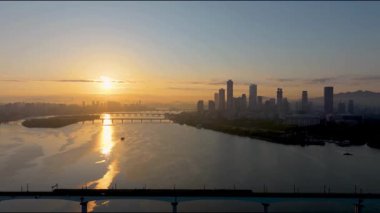 Sakin bir nehre yansıyan altın ışıkla modern bir şehrin ufuk çizgisinde güzel bir gün doğumu. Şafak vakti şehir manzarası büyümeyi, yeni başlangıçları ve metropol yaşamını simgeliyor..