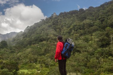 Sırt çantalı genç adam mavi gökyüzü olan sisli bir dağın tepesinde bir anıtı izliyor.