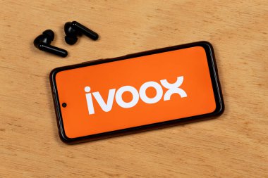 Bir akıllı telefon ekranının makro fotoğrafı ahşap arka planda kablosuz kulaklıklı Ivoox uygulaması beyaz logoyu gösteriyor.