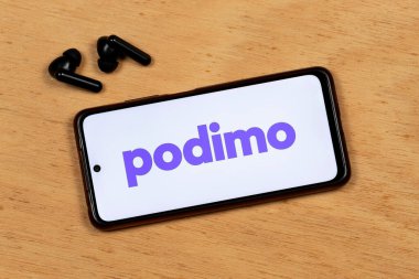 Akıllı telefon ekranının makro fotoğrafı ahşap arka planda kablosuz kulaklıkla Podimo platformu menekşe logosunu gösteriyor.