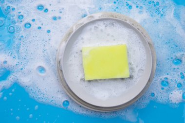 Bulaşıkları ıslak mavi arka planda sabunlu köpükle yıkıyorum..