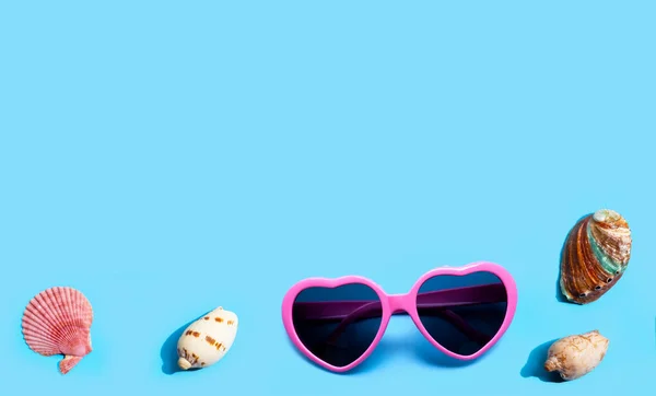 stock image Pink heart shaped sunglasses with exotic sea shells on blue background. Summer background concept