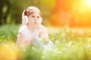 Tatlı küçük kız sonbahar parkında müzik dinliyor. Aile açık hava yaşam tarzı. Kulaklıklı mutlu küçük çocuk çimlerin üzerinde oturuyor. Sonbaharda güzellik doğası. Çocukluk mutluluğu. Çocuk günü