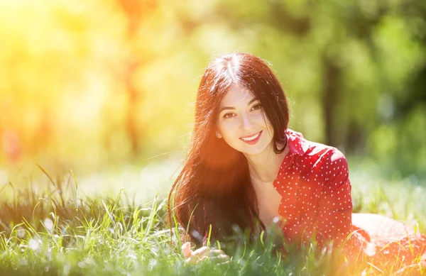 公園でリラックスした赤いドレスの若いかわいい女性 カラフルな背景を持つ美しさの自然シーン 夏の季節に木 アウトドアライフスタイル 幸せな笑顔の女性は緑の草の上に横たわっていた — ストック写真