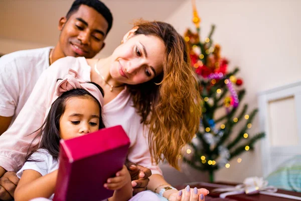 Mezcla de raza feliz familia sentado en la cama y dando una gran caja roja regalo de Navidad en la víspera de la mañana — Foto de Stock