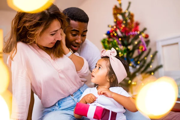 Mezcla de raza feliz familia sentado en la cama y dando una gran caja roja regalo de Navidad en la víspera de la mañana — Foto de Stock