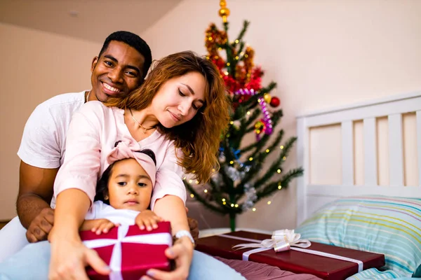 混合レース幸せな家族はベッドの上に座って、前夜に大きな赤い箱のクリスマスプレゼントを与えます — ストック写真