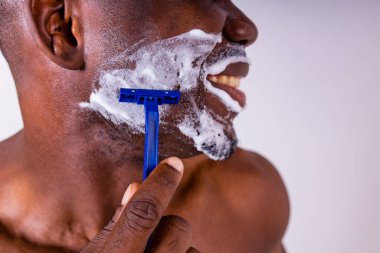 Yüzünde tıraş köpüğü ve jileti olan Latin bir adam. Stüdyo beyaz, arka planı izole ediyor.