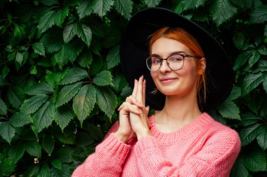 Açık havada gülümseyen kızıl pembe kazak ve yeşil yapraklı siyah şapka giyer.