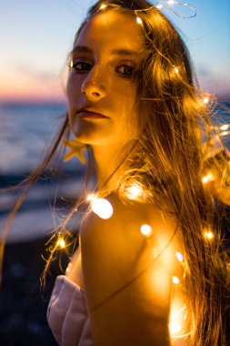 Korseli uzun saçlı kadın göğsünde kabuklar olan deniz kıyısında alacakaranlıkta uyuşuk görünüyor.