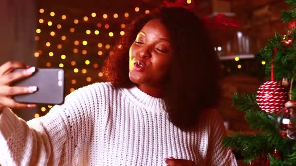 Афроамериканська жінка в білому светрі і розі оленя робить селфі і розмовляє на фоні домашнього вогню — стокове відео